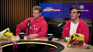 Dwóch mężczyzn w czerwonych bluzach z kapturem siedzi przy stole w studiu telewizyjnym, przed nimi mikrofony, kubki i bukiety kwiatów z medalami.