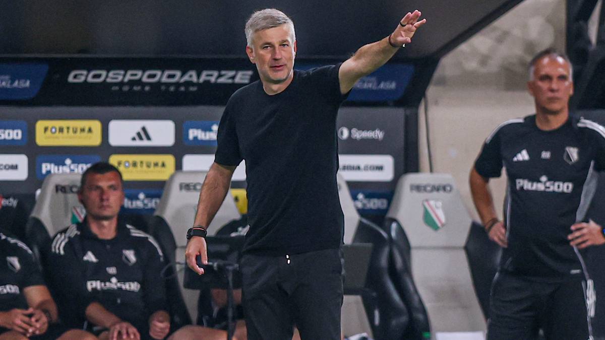 Trener piłkarski w czarnym stroju, stojący na boisku, z uniesioną prawą ręką, gestykulujący. Obok niego siedzą na ławce rezerwowi.