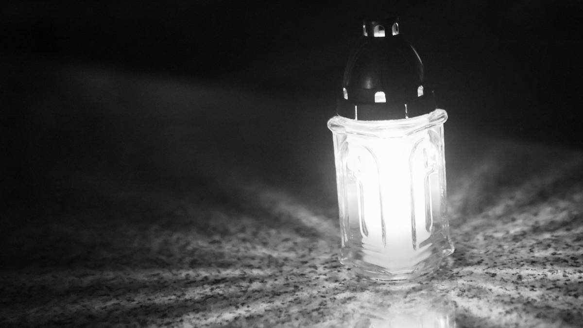 Zbliżenie na płonący znicz na ciemnym tle cmentarza nocą. Światło świecy jest jasne i rozchodzi się promienisto.