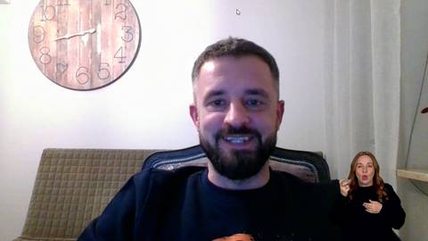 Artur Gac uśmiecha się do kamery. W tle za nim znajduje się zegar i kanapa. Po prawej stronie widoczna jest tłumaczka języka migowego.