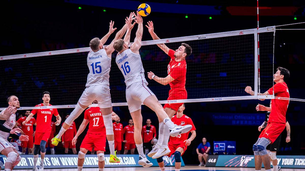 Sześciu siatkarzy w akcji przy siatce. Dwóch zawodników w białych strojach blokuje atak wykonany przez dwóch zawodników w czerwonych strojach. Jeden z białych graczy wyskakuje do bloku, a drugi próbuje go zatrzymać. Jeden z czerwonych graczy próbuje przebić się przez blok. Trzeci czerwony gracz ogląda akcję z boku, a czwarty stoi na drugim końcu boiska.