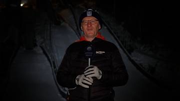 Mężczyzna w czapce, okularach i rękawiczkach trzyma mikrofon w ciemnej scenerii.