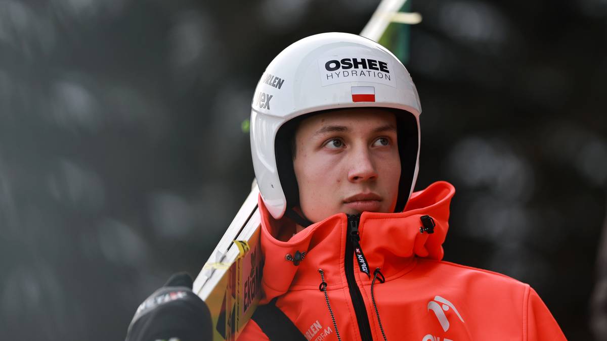 Skoczek narciarski w białym kasku i pomarańczowej kurtce z nartami na ramieniu.