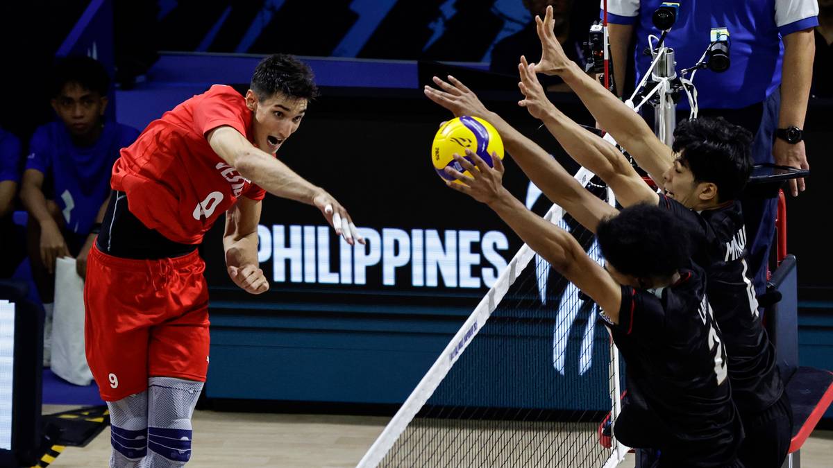 Siatkarz w czerwonej koszulce serwuje piłkę, podczas gdy trzech przeciwników w czarnych strojach próbuje ją zablokować.