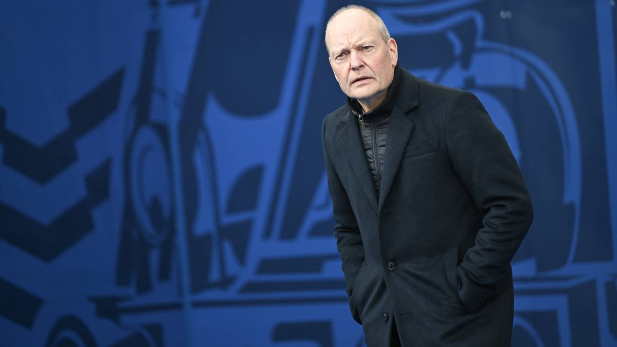 Starszy mężczyzna w ciemnym płaszczu, z łysą głową, stoi na tle niebieskiej ściany z czarnymi, geometrycznymi wzorami. Ma poważny wyraz twarzy.