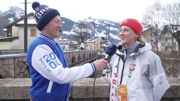 Mężczyzna w czapce zimowej i kurtce z napisem "20 born in" przeprowadza wywiad z kobietą w czerwonej czapce i kurtce zimowej, trzymając mikrofon.