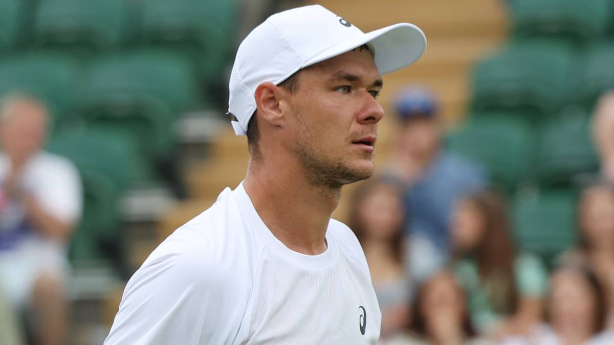 Portret tenisisty Kamila Majchrzaka w białej czapce i koszulce, patrzącego w bok z wyrazem koncentracji na twarzy. W tle widać zielone trybuny i rozmazane sylwetki widzów.