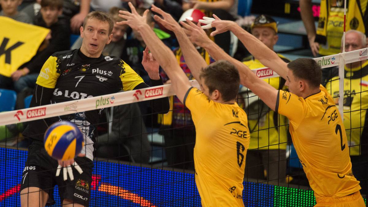 Siatkarze w żółtych strojach blokują piłkę nad siatką, zawodnik w czarnym stroju z numerem 7 po lewej stronie przygotowuje się do zagrania.