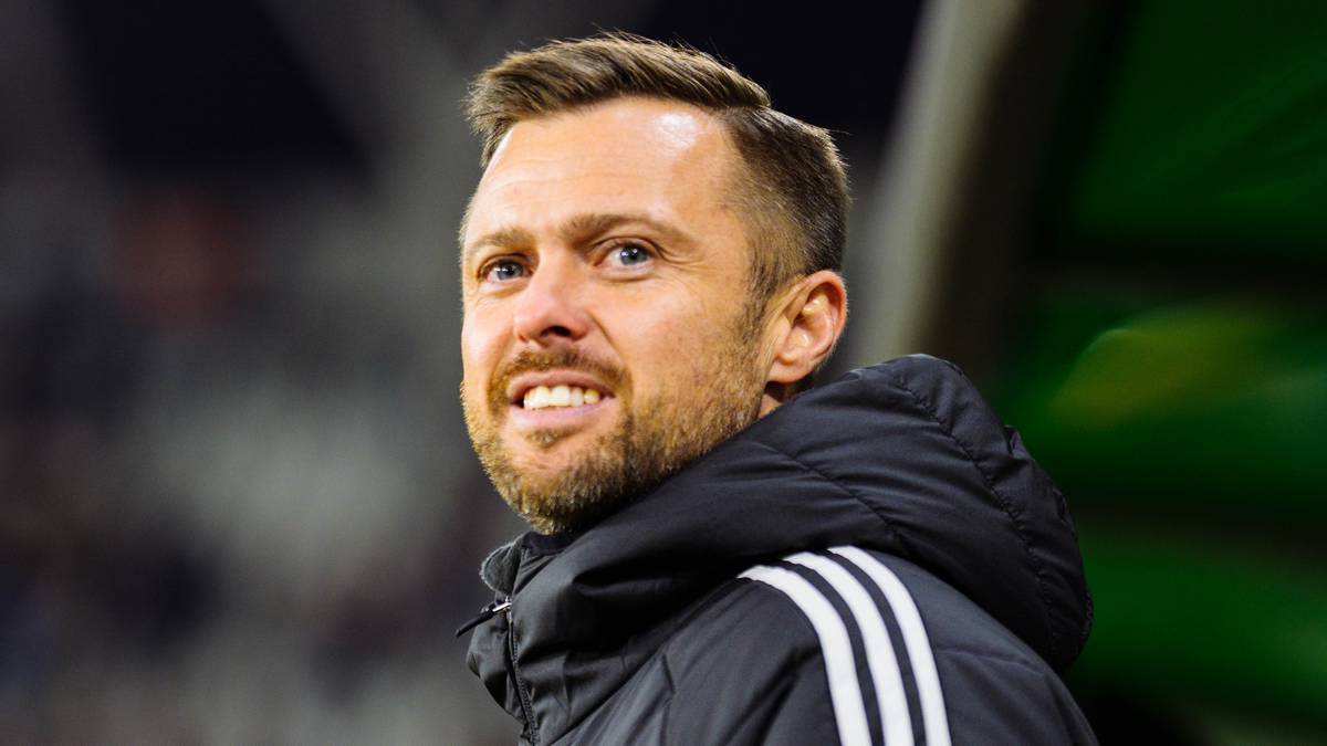 Mężczyzna w czarnej kurtce z białymi pasami na ramieniu, uśmiecha się.