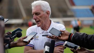 Starszy mężczyzna o siwych włosach, ubrany w białą koszulkę polo, otoczony przez ręce trzymające mikrofony i telefony kom&oacute;rkowe, udziela wywiadu.