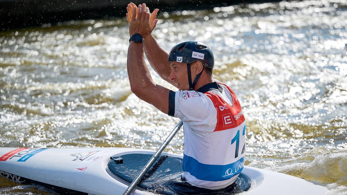 Kajakarz w kasku i koszulce sportowej z numerem 11, wiosłuje na wodzie, z rękami uniesionymi w g&oacute;rę.