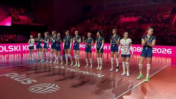 Grupa siatkarek w niebieskich strojach stoi w szeregu na boisku siatkarskim, patrząc w stronę widowni.