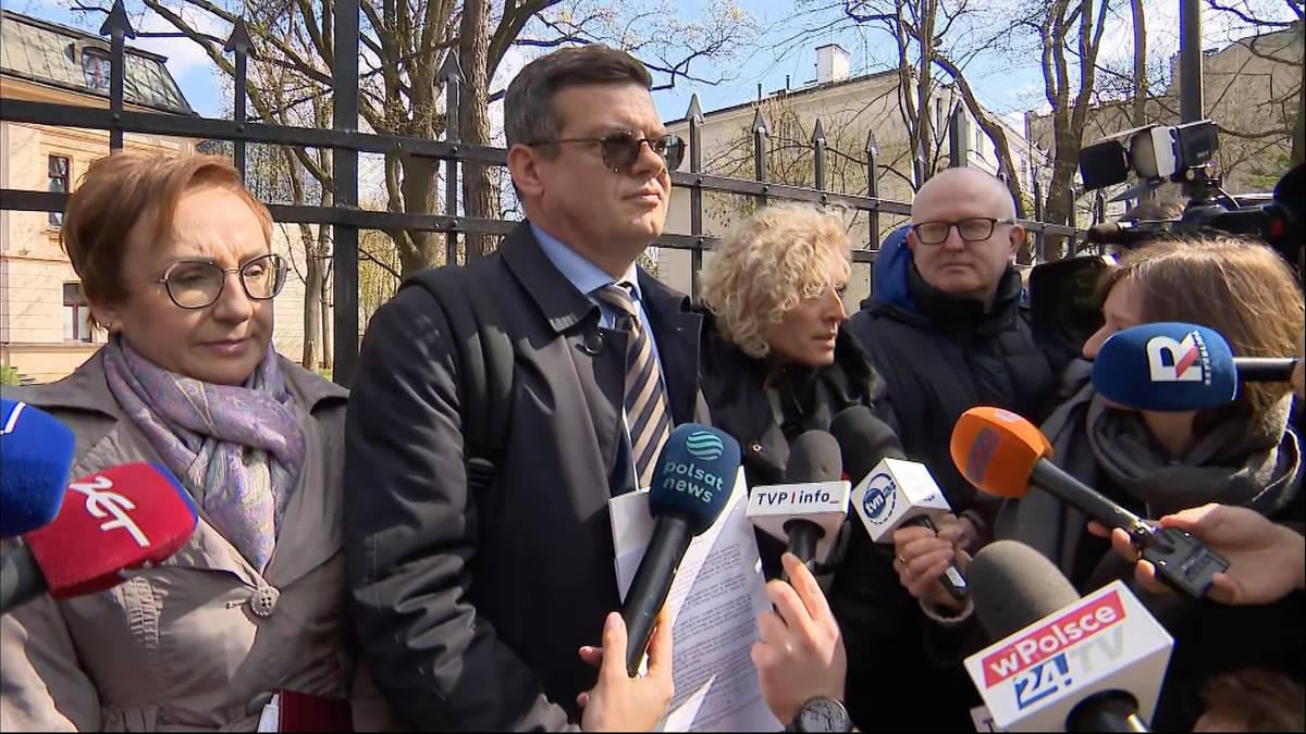 Sędzia Krystian Markiewicz przemawia do mediów, trzymając w ręku dokumenty, otoczony przez innych sędziów i dziennikarzy z mikrofonami.