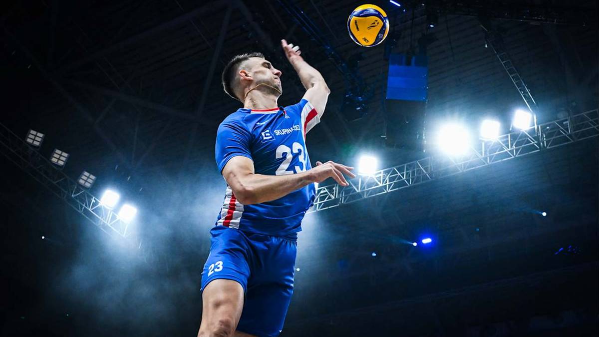 Siatkarz w niebieskiej koszulce z numerem 23 serwuje piłkę do siatkówki w hali sportowej.