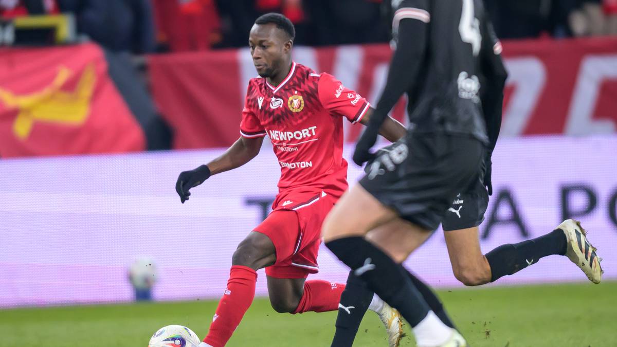 Piłkarz w czerwonej koszulce z numerem 7 prowadzący piłkę, ścigany przez innego zawodnika.