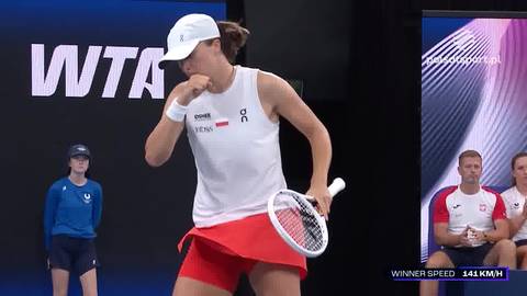 Iga Świątek, polska tenisistka, w białej koszulce i czerwonej sp&oacute;dniczce, z białą czapką na głowie, cieszy się z udanego zagrania podczas meczu tenisowego.