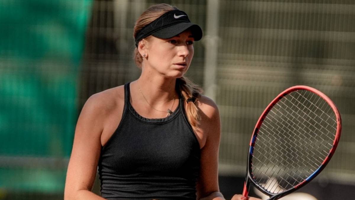 Kobieta tenisistka w czarnym topie i czapce z daszkiem z logo, trzymająca rakietę tenisową.