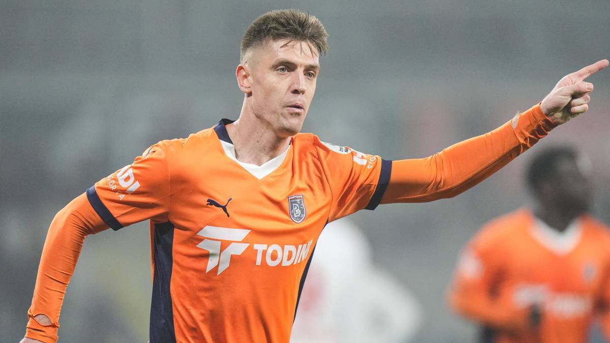 Krzysztof Piątek w pomarańczowej koszulce piłkarskiej z białymi elementami, wskazujący prawą ręką w bok.