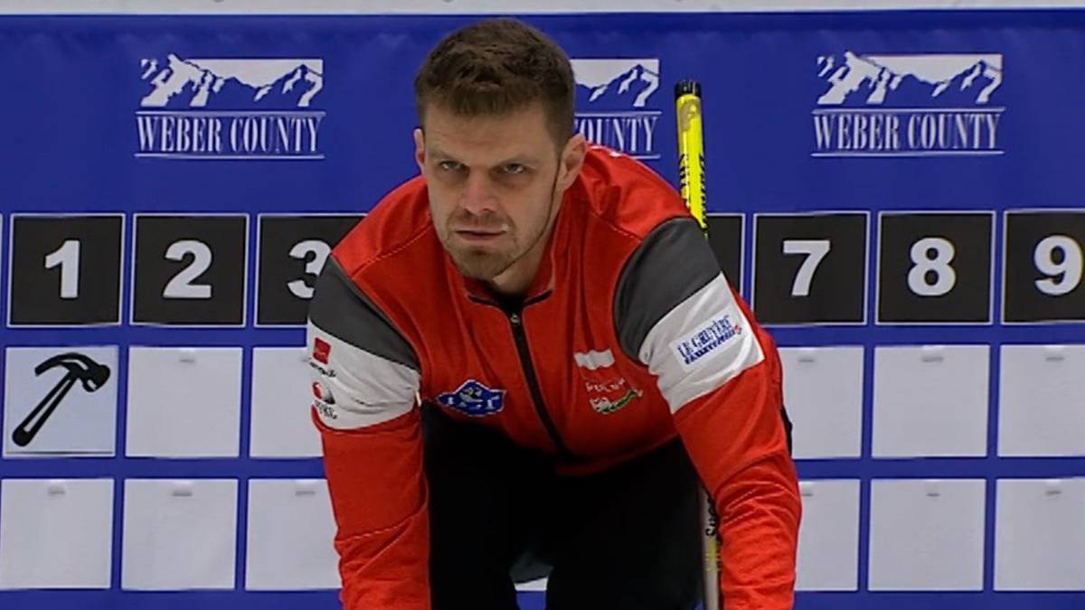 Curler w czerwonej kurtce sportowej kuca na lodowisku, przygotowując się do rzutu kamieniem.