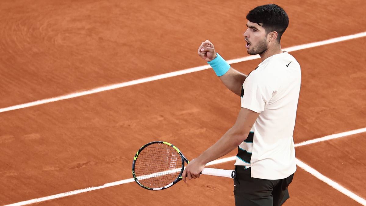 Carlos Alcaraz, tenisista w białej koszulce i czarnych spodenkach, świętuje zwycięstwo na ceglanej nawierzchni kortu tenisowego Rolanda Garrosa.