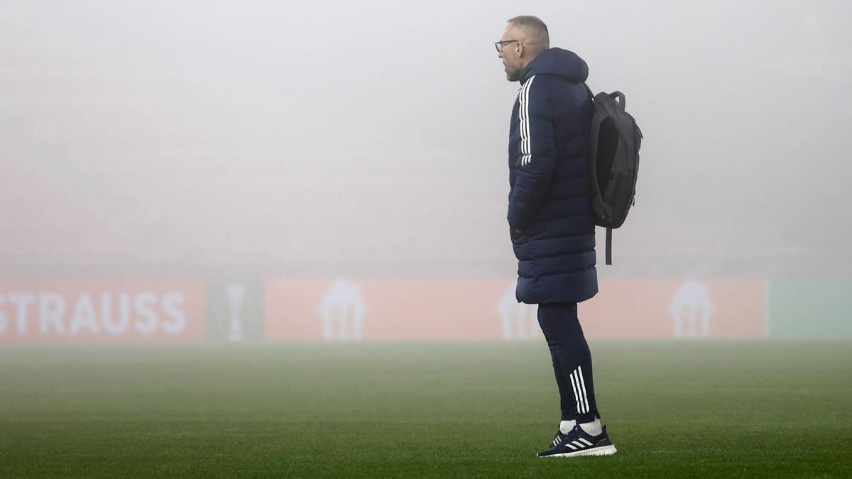 Trener Marek Papszun w ciemnej kurtce i z plecakiem stoi na boisku piłkarskim pokrytym mgłą.