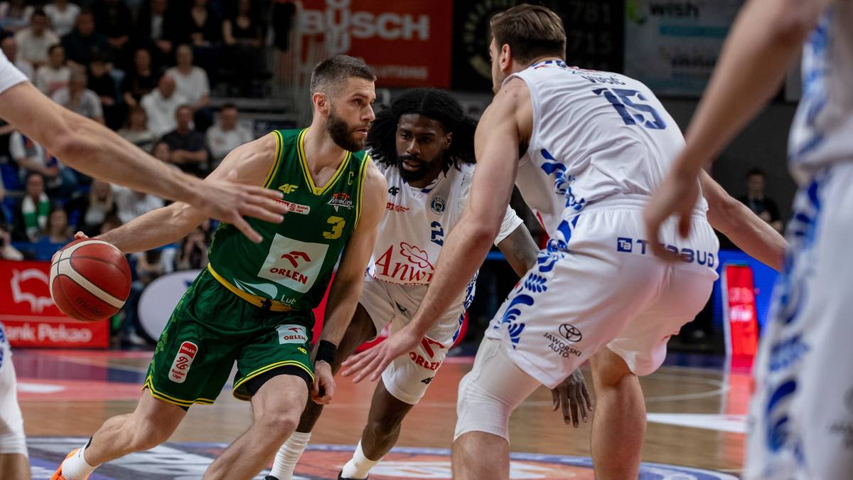 Gracz w zielonej koszulce z numerem 3 drybluje piłką do koszyk&oacute;wki, otoczony przez dw&oacute;ch graczy w białych koszulkach. Jeden z nich, z numerem 15, pr&oacute;buje go zatrzymać.