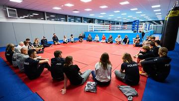 Grupa młodych ludzi siedzi w kręgu na macie, słuchając osoby stojącej pośrodku.