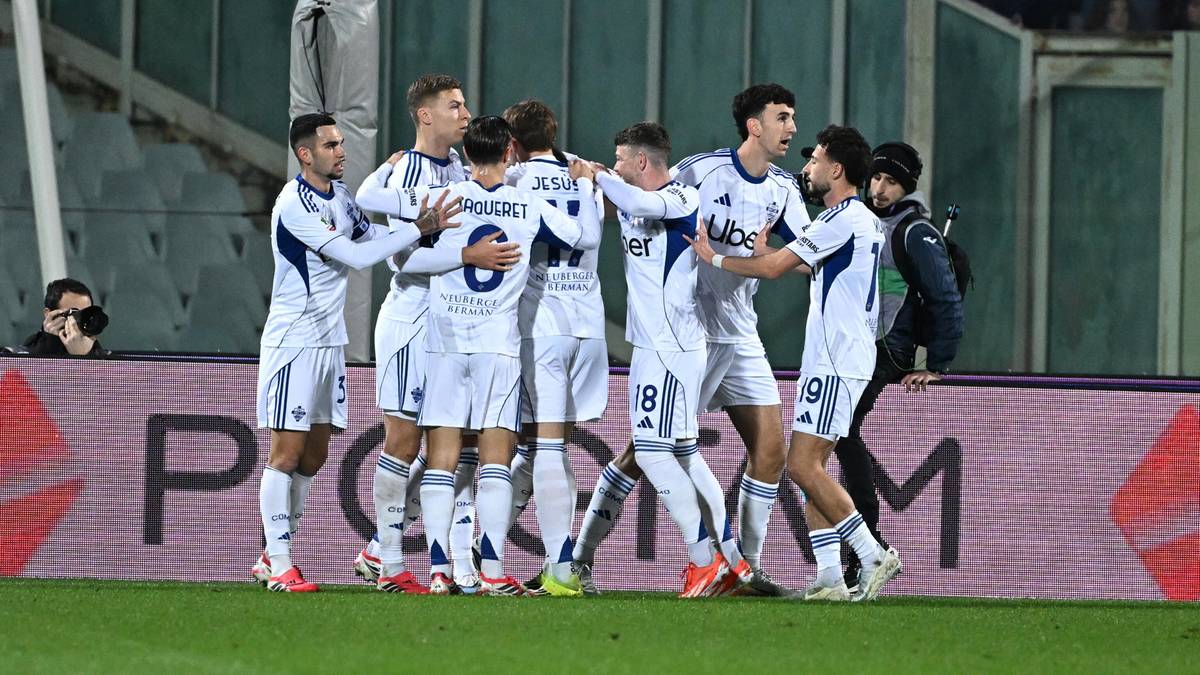 Drużyna piłkarska świętuje bramkę, gracze w białych strojach z niebieskimi elementami są zgrupowani na boisku.