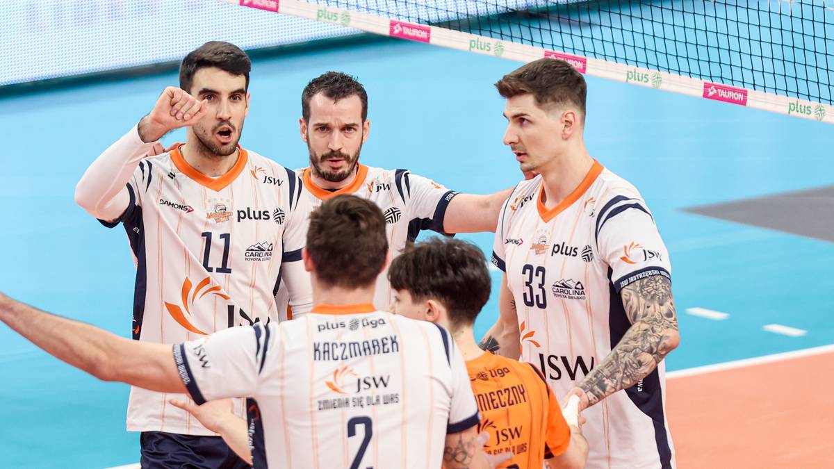 Czterech siatkarzy w białych strojach z pomarańczowymi detalami stoi na boisku siatkarskim, rozmawiając ze sobą.