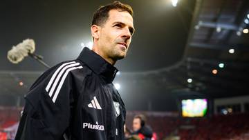 Trener Legii Warszawa w kurtce sportowej z logo Adidas, patrzący w bok. W tle stadion i kibice.