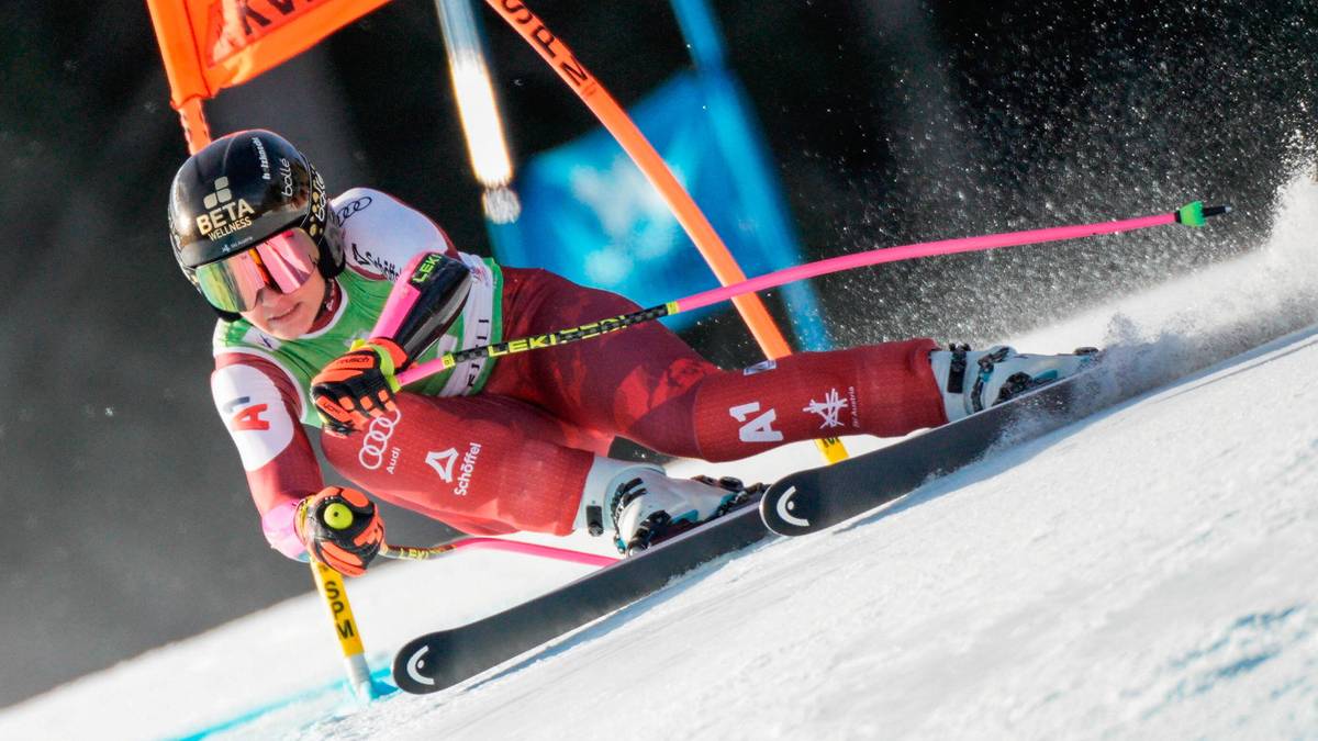 Kobieta narciarka w czerwonym stroju i białym kasku zjeżdża po zaśnieżonym stoku, narzucając narty i trzymając kije narciarskie.