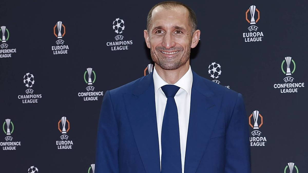Giorgio Chiellini w niebieskim garniturze i białej koszuli z niebieskim krawatem, uśmiechnięty, na tle ściany z logotypami Ligi Mistrzów, Ligi Europy i Ligi Konferencji.