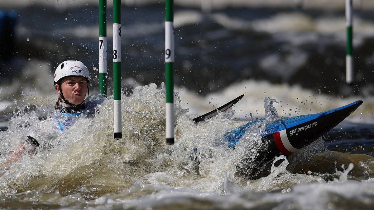 Zawodniczka w kasku i okularach pływa w rozbryzgach wody obok wywróconego kajaka.