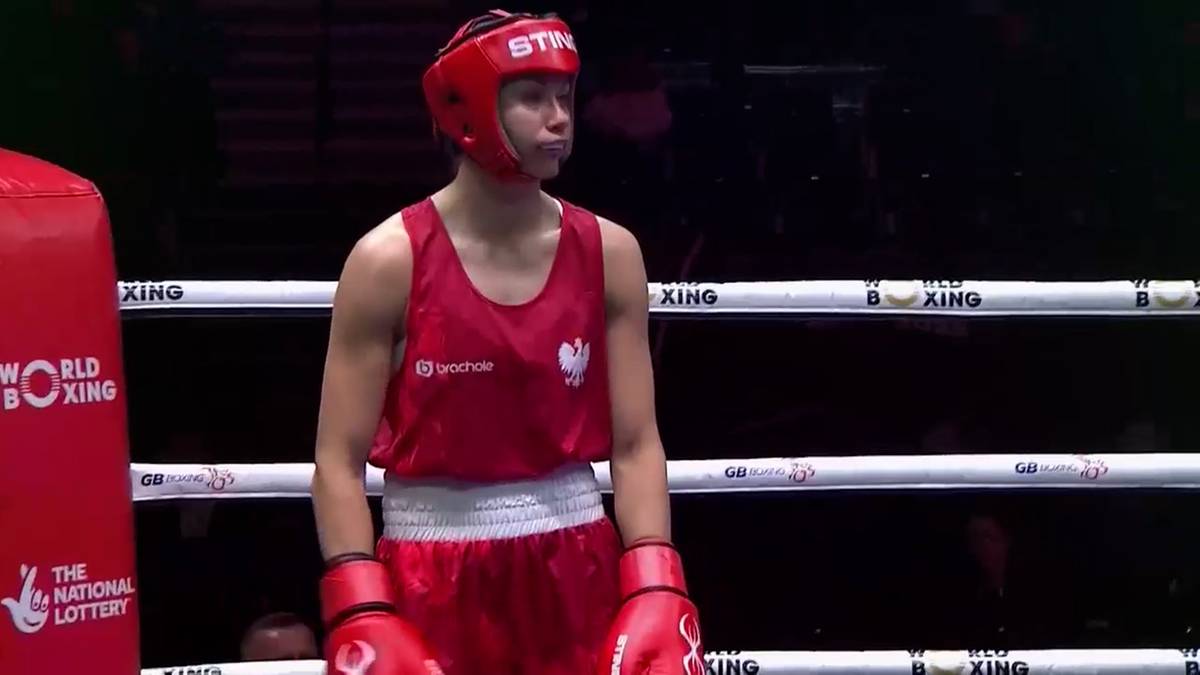 Bokserka w czerwonym stroju sportowym i kasku stoi na ringu bokserskim, patrząc w bok.