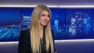 Kobieta z blond włosami, ubrana w ciemną marynarkę, stoi na tle nocnej panoramy miasta.