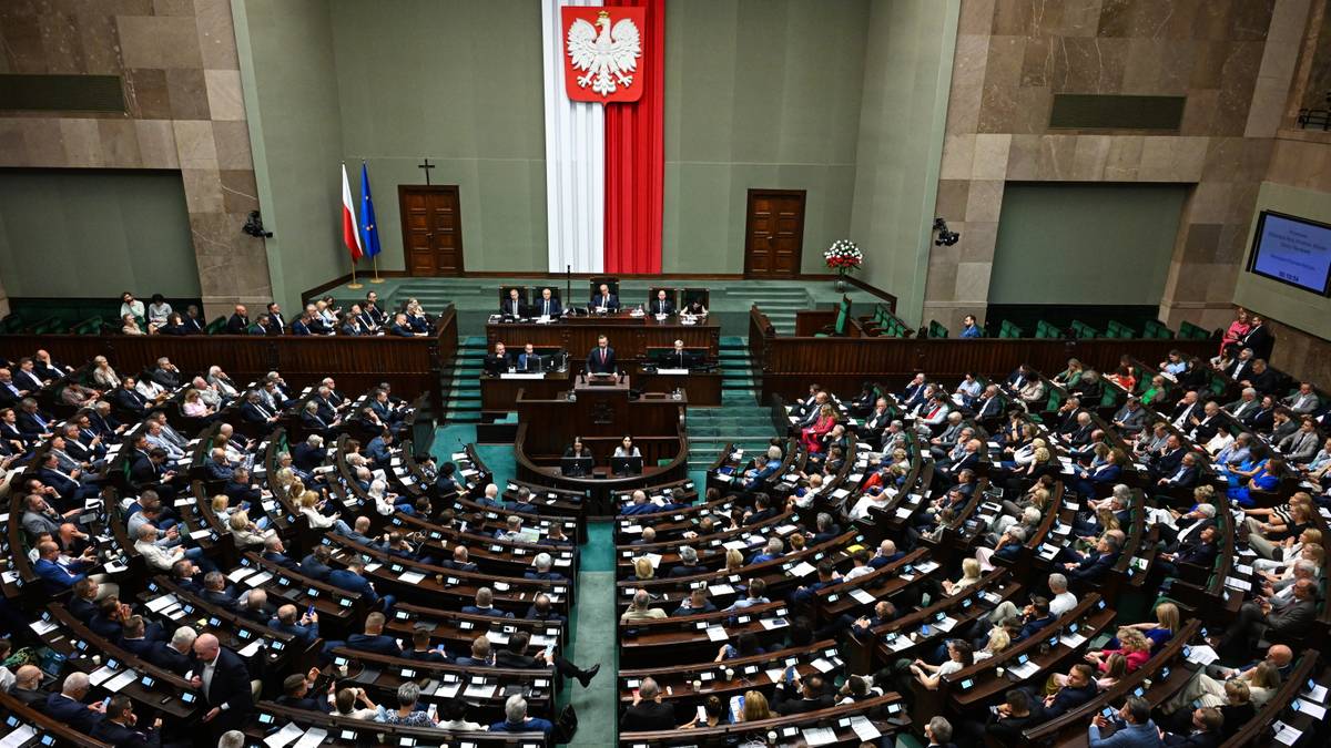 Widok na salę plenarną Sejmu Rzeczypospolitej Polskiej z posłami siedzącymi w ławach. Na ścianie wisi godło Polski oraz flagi Polski i Unii Europejskiej.