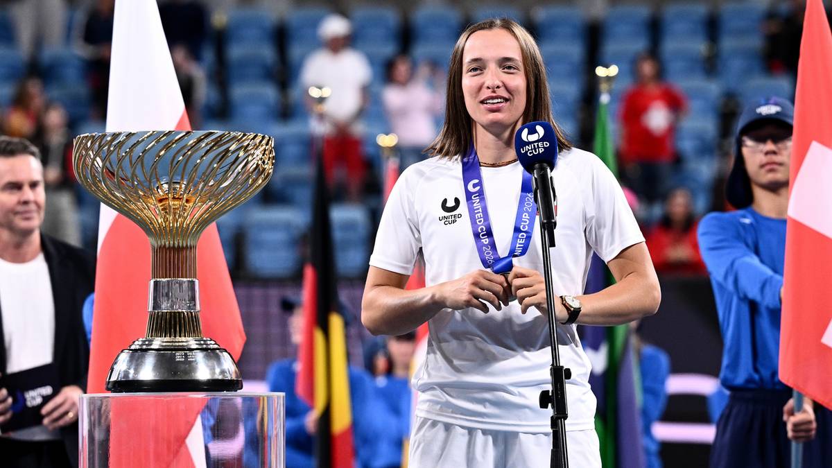 Iga Świątek w stroju tenisowym z medalem na szyi, stoi przy mikrofonie obok pucharu i flag.