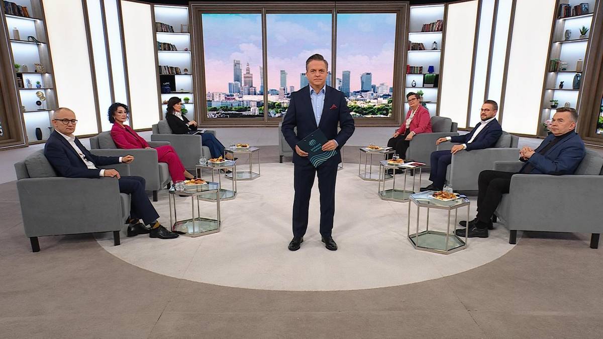 Moderator w garniturze stoi pośrodku studia telewizyjnego, trzymając teczkę. Wok&oacute;ł niego, siedząc na kanapach, znajduje się siedmiu uczestnik&oacute;w dyskusji.