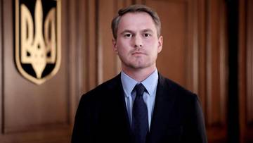 Mężczyzna w garniturze na tle drewnianej ściany z herbem Ukrainy.