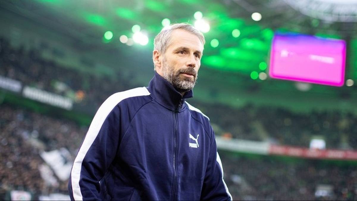 Mężczyzna w średnim wieku w sportowej kurtce z kołnierzem, stojący na tle stadionu piłkarskiego z zielonymi światłami i dużym ekranem.