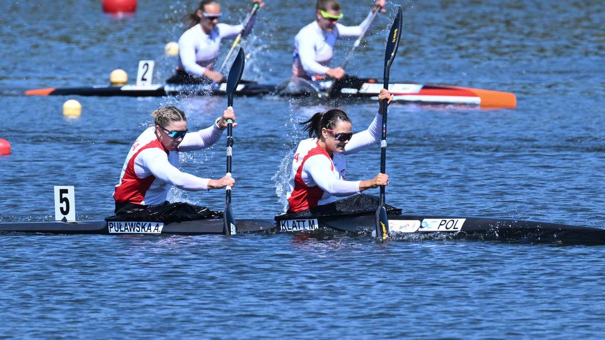 Dwie kobiety w kajaku, w białych koszulkach z czerwonymi wstawkami i numerem 5, wiosłują podczas wyścigu kajakowego.