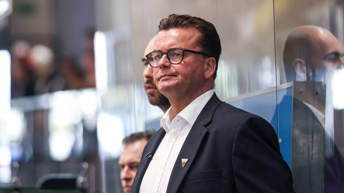 Mężczyzna w okularach i garniturze, patrzy w bok z poważną miną. Za nim widać innych mężczyzn i nieostre tło.