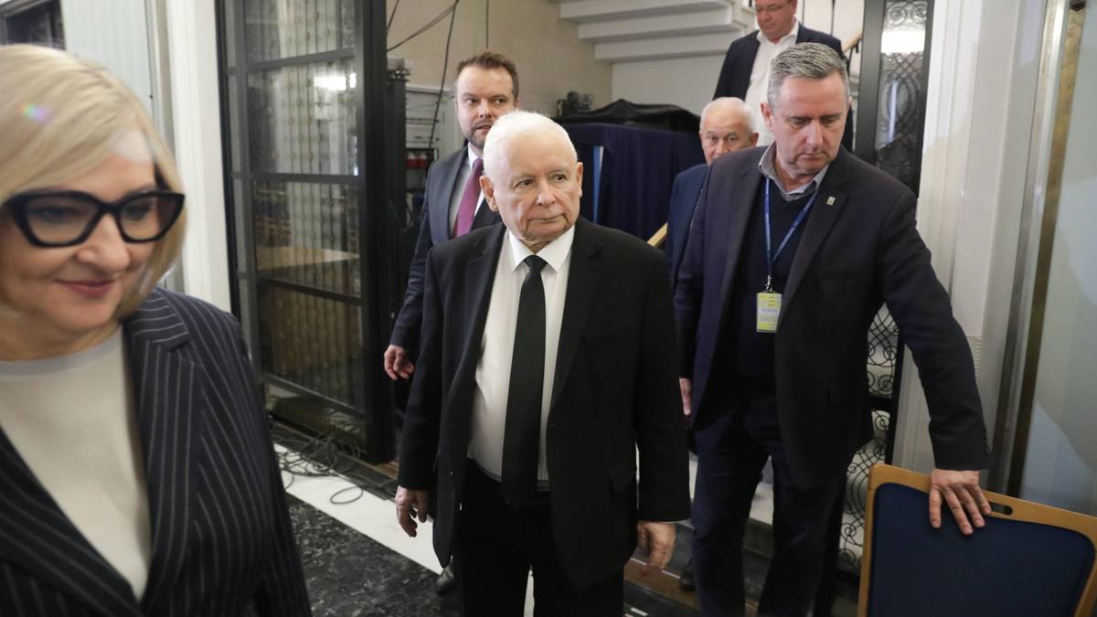 Jarosław Kaczyński w garniturze i krawacie w otoczeniu innych mężczyzn w budynku.