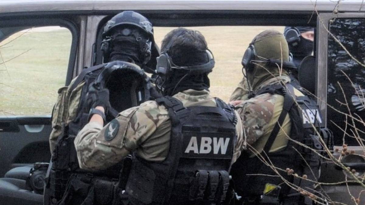 Trzech funkcjonariuszy Agencji Bezpieczeństwa Wewnętrznego w pełnym umundurowaniu i wyposażeniu taktycznym, z widocznymi kamizelkami z napisem "ABW". Jeden z nich stoi tyłem, dwaj pozostali są odwróceni bokiem, rozmawiają ze sobą.
