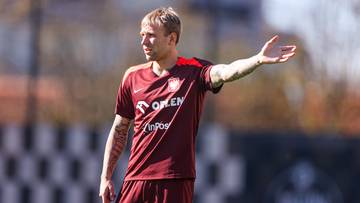 Piłkarz w czerwonej koszulce z białym logo i napisami wykonuje gest prawą ręką.