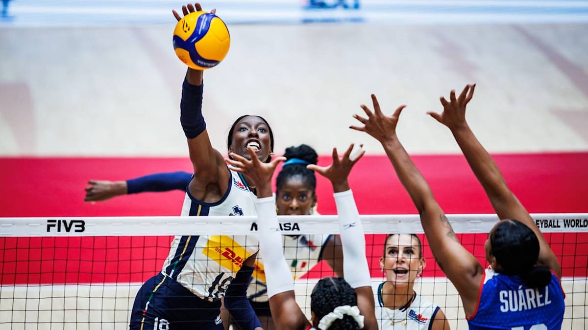 Siatkarka w czarnej koszulce z żółtymi elementami wykonuje wysoki skok do ataku, uderzając piłkę ręką w górę. Jej oczy są skupione na piłce, a twarz wyraża determinację. Za nią, po drugiej stronie siatki, inne zawodniczki próbują zablokować atak.