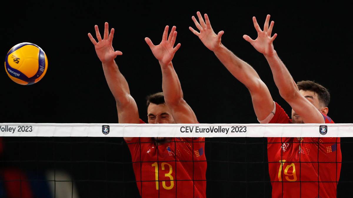 Siatkarz wykonuje blok w meczu siatkówki, piłka znajduje się w powietrzu obok jego dłoni. Na drugim planie widać dwóch innych zawodników z rękami uniesionymi do góry.