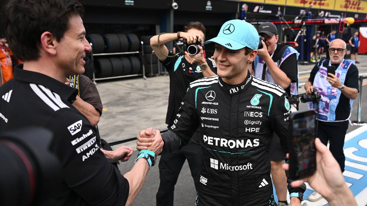 Dw&oacute;ch kierowc&oacute;w Formuły 1 uściskuje sobie dłonie na tle toru wyścigowego.