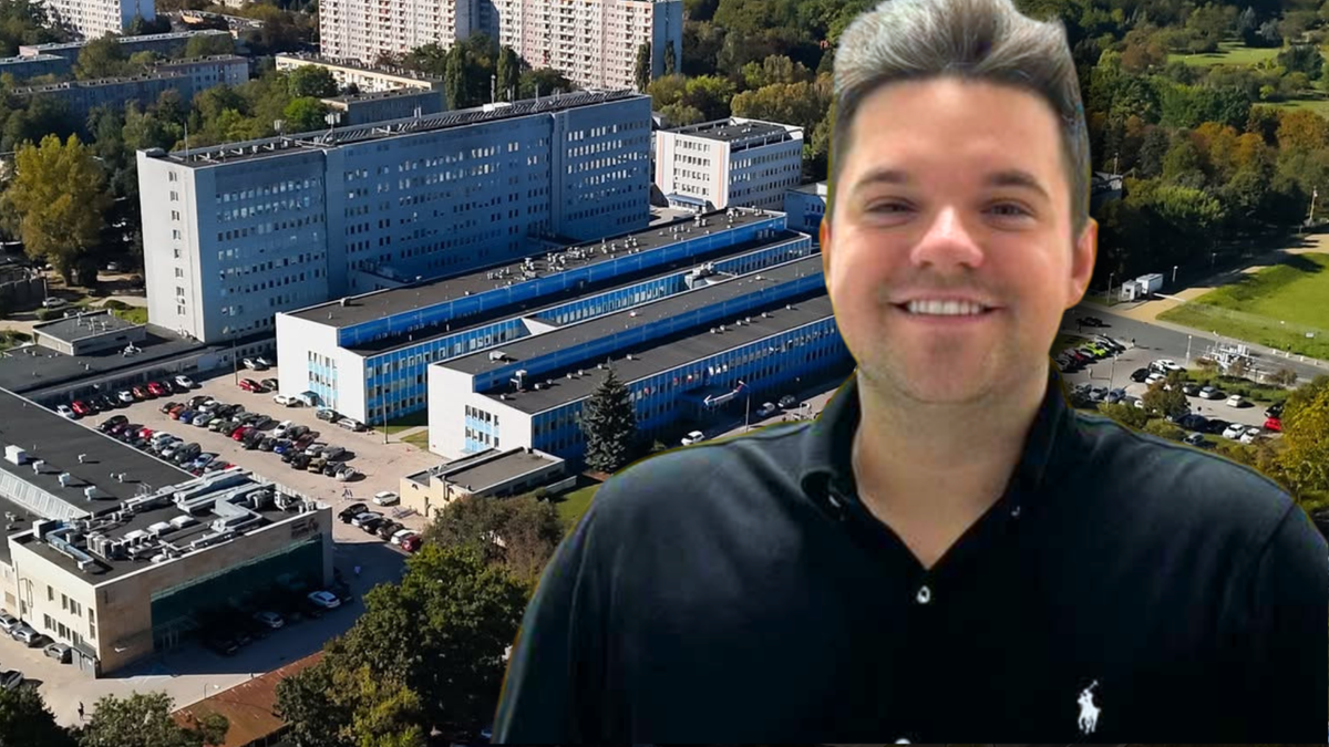 Mężczyzna uśmiecha się na tle dużego kompleksu budynków szpitalnych.