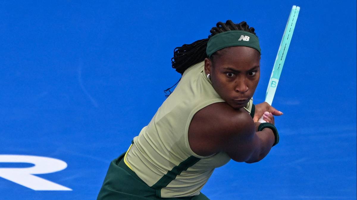 Tenisistka w ciemnozielonej opasce na głowie i żółtej koszulce z krótkim rękawem wykonuje zamach rakietą tenisową na tle niebieskiego boiska.